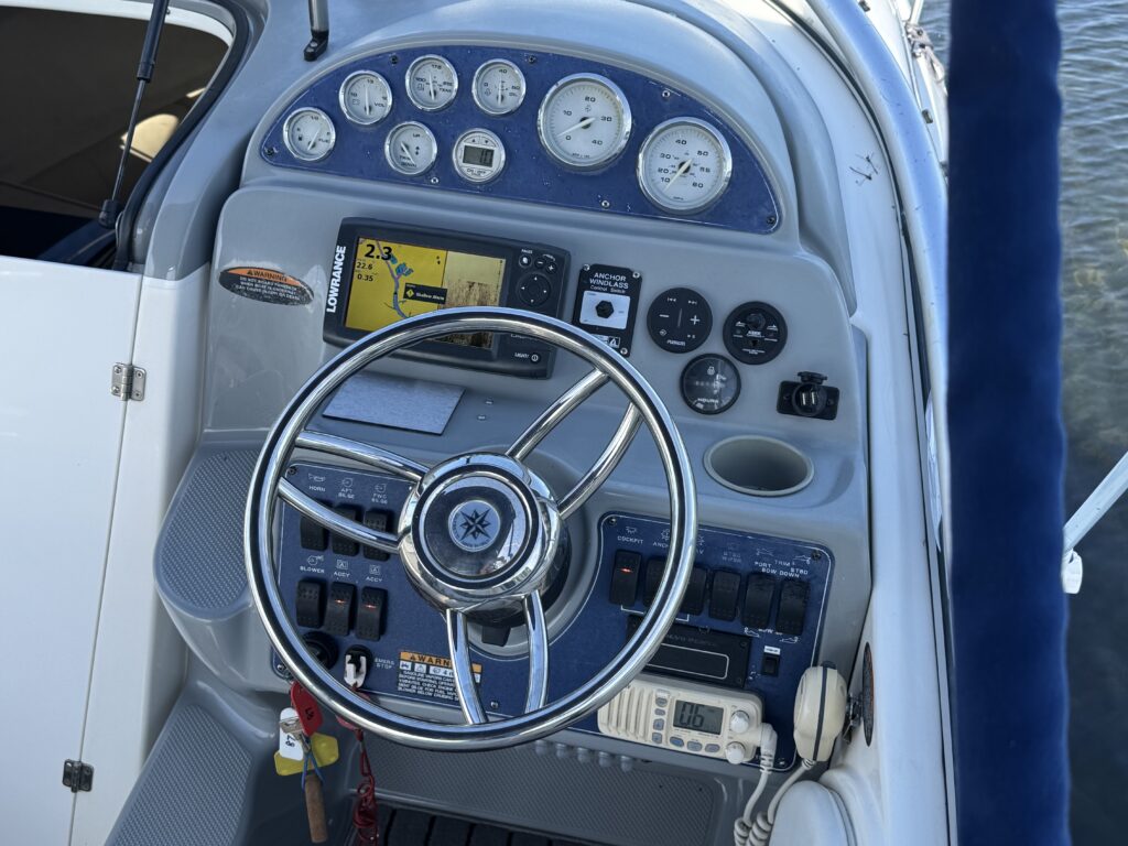 The control console of a boat featuring a silver steering wheel, various gauges for speed and fuel, a GPS screen, and multiple switches for equipment. A communication device is visible on the side, with a clean gray and blue interior.