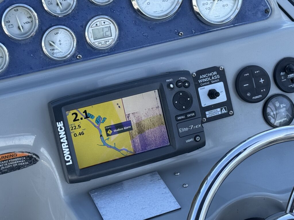 A Lowrance Elite-7 fish finder mounted on a boat's dashboard displays navigation data, including depth readings and a color map of the area. The dashboard features various gauges and controls adjacent to the monitor.