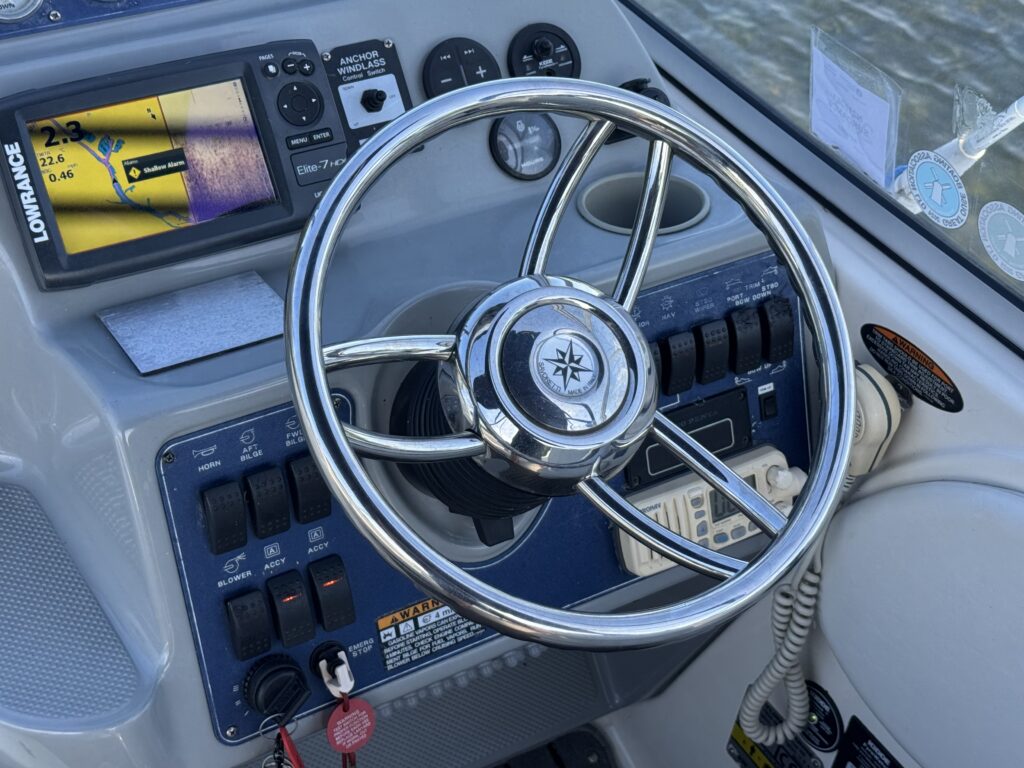The image shows a close-up view of a boat's control panel with a shiny steering wheel in the center. To the left, there is a digital display and various switches and knobs, while the background features water reflecting natural light.