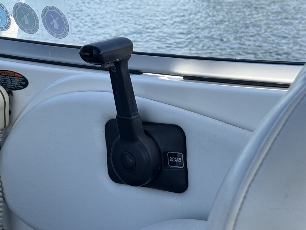A black manual adjustment lever positioned beside a white upholstered boat seat. The background shows a body of water, and circular stickers are visible on the window above the lever.