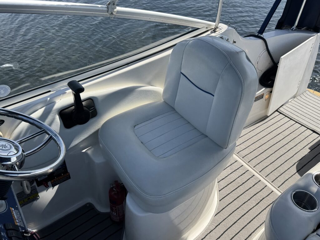 A white upholstered captain's seat is positioned on a boat, with a steering wheel and control lever visible on the left. The boat's textured flooring and water surface are seen in the background.
