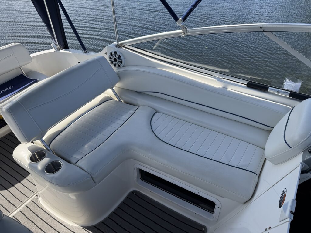 A white and gray boat seat with a curved backrest is positioned in a marine setting. The seat features integrated cup holders and is surrounded by blue canvas and a view of the water in the background.