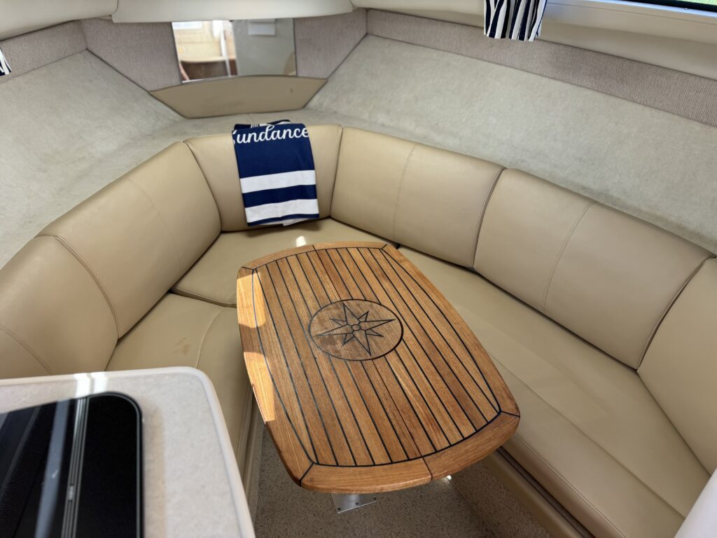 A beige leather seating area inside a boat, featuring a wooden table with a compass design in the center. A striped towel with the word "Sundance" is draped over the back of the seating. The walls are light-colored, and there are large windows allowing natural light.