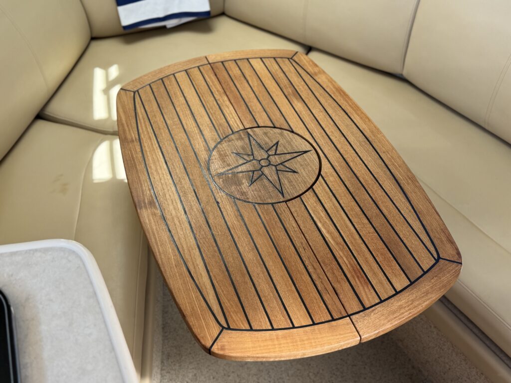 A wooden table with a compass rose design, featuring horizontal and vertical stripes, positioned on a light-colored seating area. The table is surrounded by a beige leather sofa, and sunlight is reflecting off the surface.