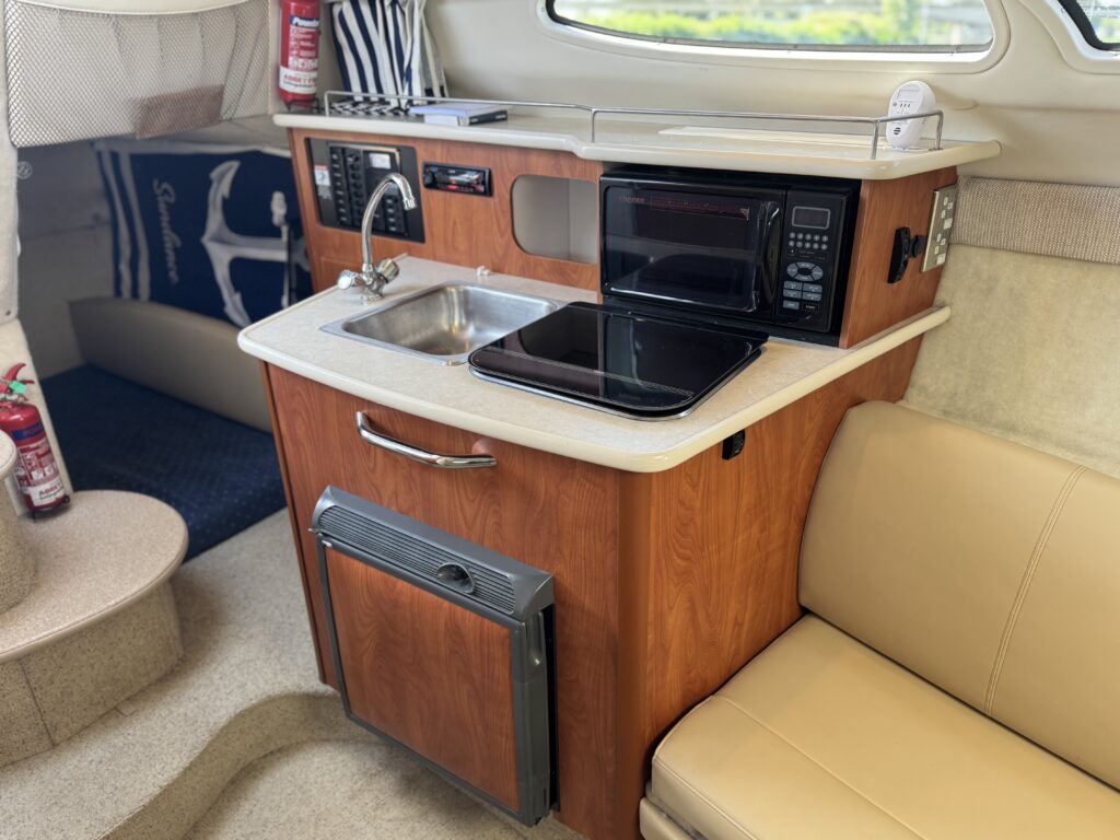 A compact kitchen setup inside a boat featuring a wooden cabinet with a sink, a microwave, and a small refrigerator. The countertop has a black stovetop, and the area includes a cushioned beige seat and blue carpeting.