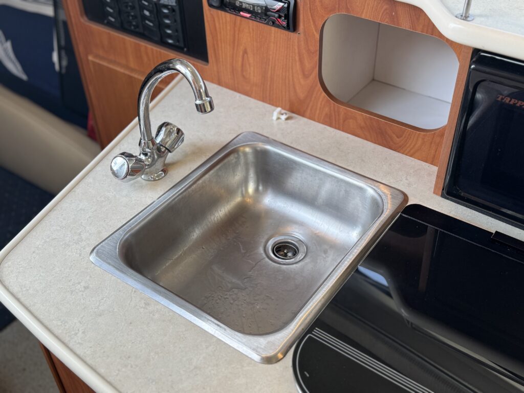 A stainless steel sink with a faucet installed on a light-colored countertop. The sink is surrounded by wooden cabinetry, and a portion of an appliance is visible on the right side. The background includes a dark blue fabric, possibly a cushion.