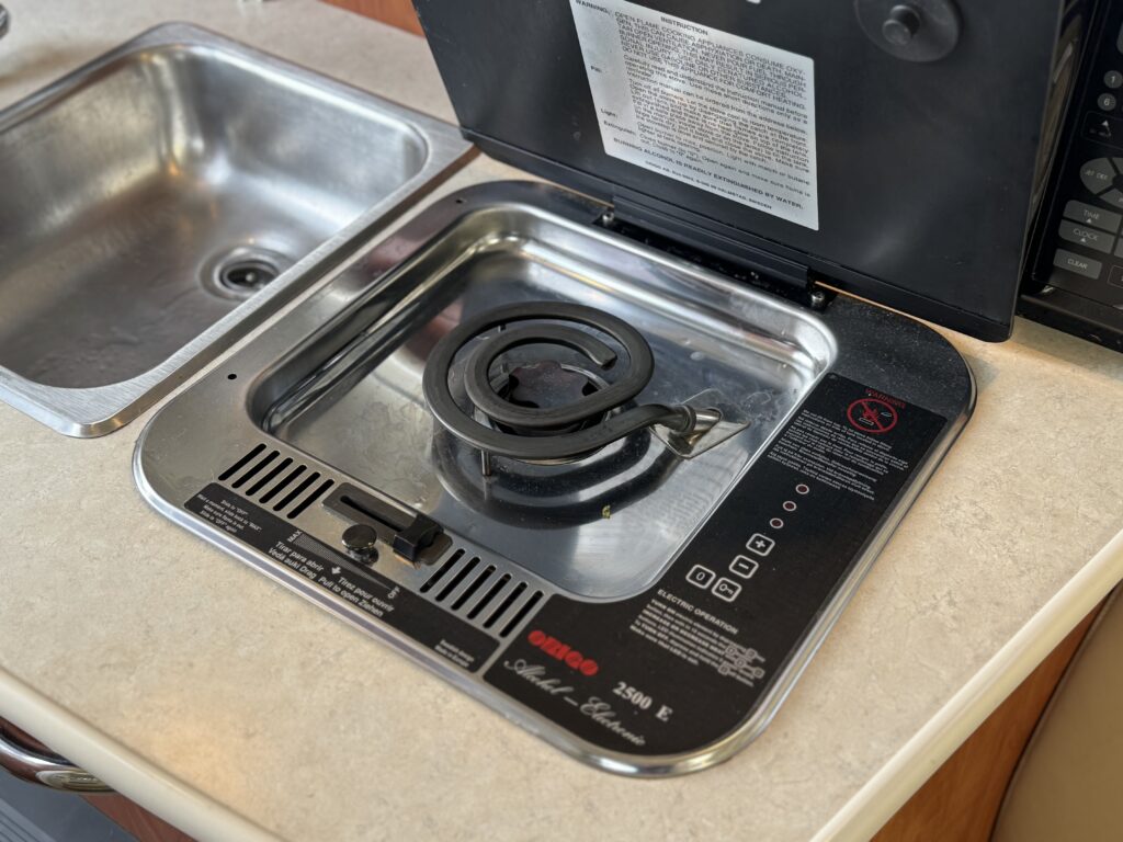 A compact stainless steel gas burner integrated into a countertop, with a round burner placed over a grid support. A nearby sink is visible, and the surface has a beige pattern. The burner has control markings for operation.