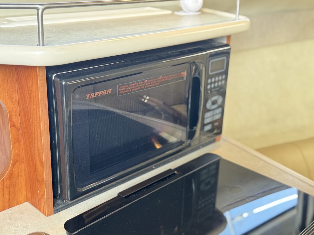 A black microwave oven is built into a wooden cabinet. The microwave features a digital display and buttons on the front and sits above a black countertop. The background includes a beige wall and a small white object on the countertop.