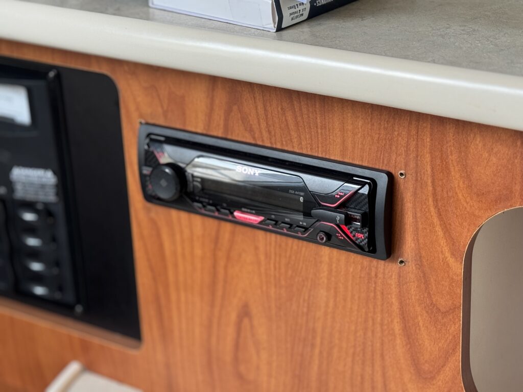 A Sony car stereo unit mounted into a wooden panel, featuring a sleek black design with red accents. To the right, a portion of an adjacent surface is visible, along with some electronic components in the background.