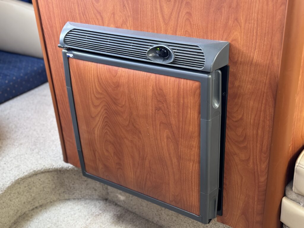 A compact refrigerator is integrated into a wooden cabinet, featuring a gray frame and a front panel that is wood-like in appearance. The background includes a soft blue upholstery and a light-colored floor, suggesting a interior space.