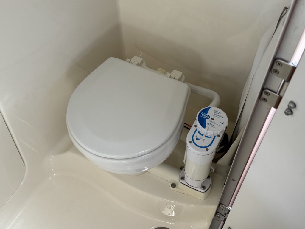 A white toilet with a closed lid is installed in a small bathroom space. Next to the toilet, a compact macerator pump with control buttons is positioned on the floor, and the walls and floor are light-colored.