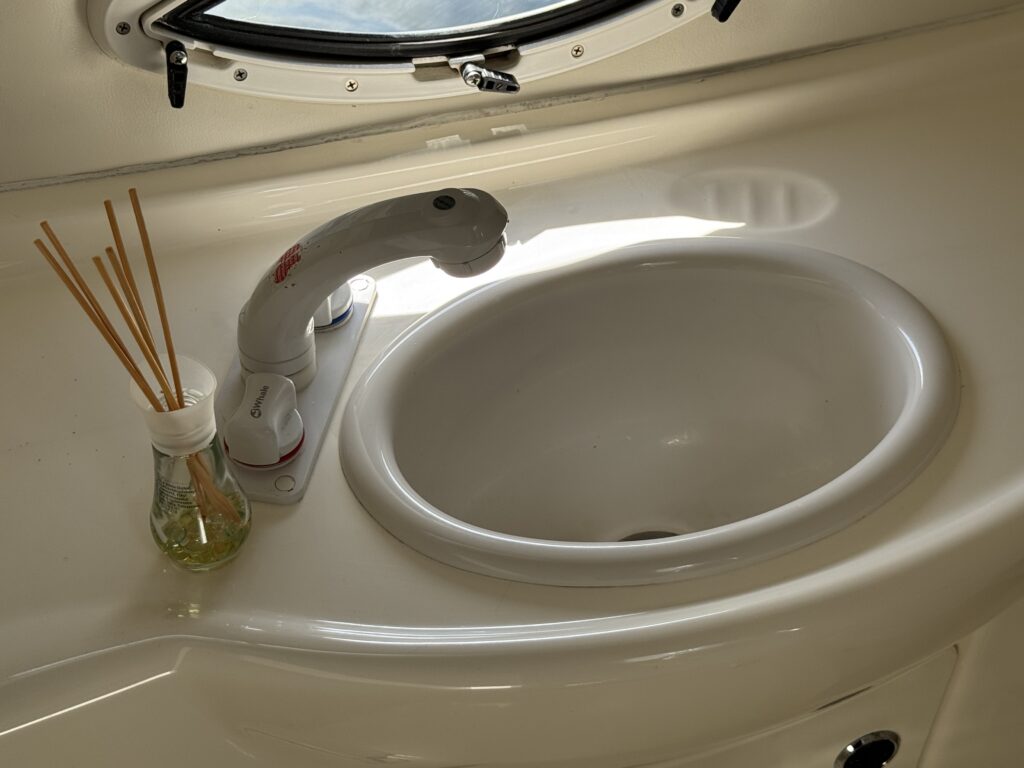A white sink with a curved basin installed in a light-colored countertop. A modern faucet with a curved design is positioned above the sink, and a glass container with reed diffusers is placed next to it.