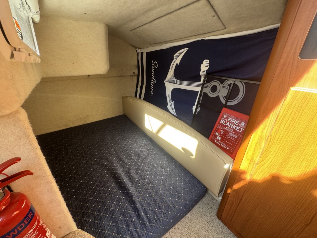 A cozy space featuring a dark blue mattress on a beige platform, next to a fire extinguisher and a safety poster for a fire blanket. A navy and white anchor-patterned blanket is draped on the wall, while natural light enters from the window.