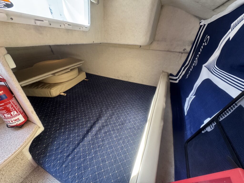 An interior shot of a small cabin featuring a blue patterned mattress on the floor, a cream-colored cushion beside it, and a white bench along one wall. A fire extinguisher is visible against the left wall, and a navy blue fabric with a logo is draped on the right side.