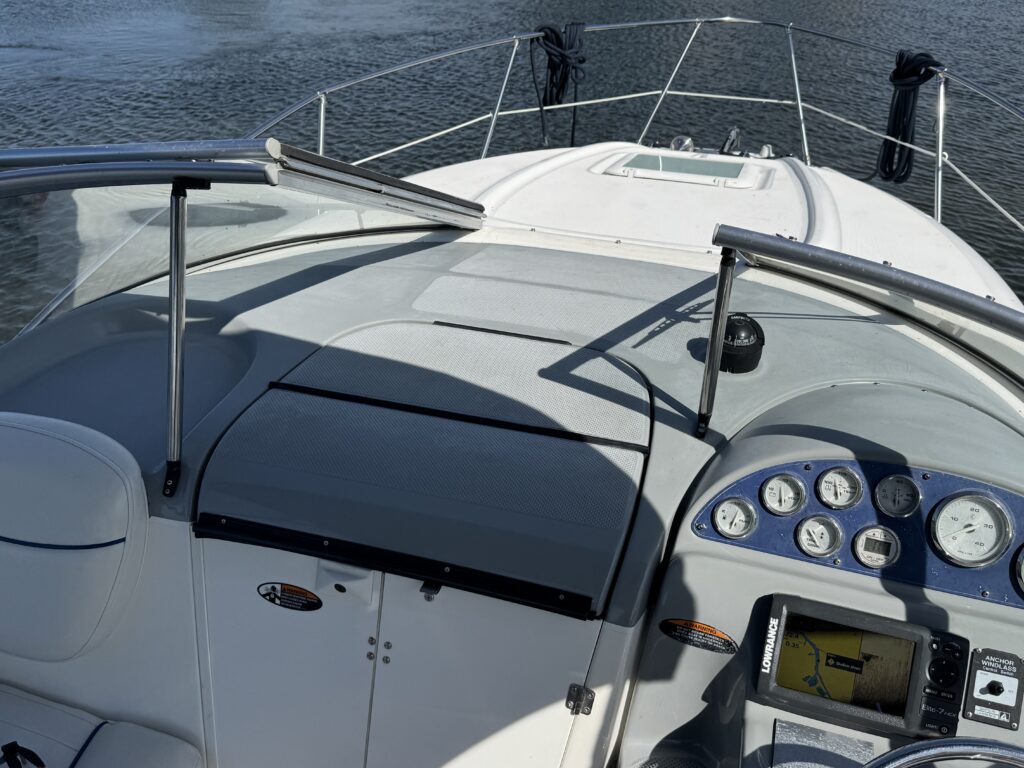 View of the dashboard of a motorboat, featuring a steering wheel, navigation instruments, and control panel. The boat's interior is light-colored, with a covered area and rails on the sides, and water visible in the background.