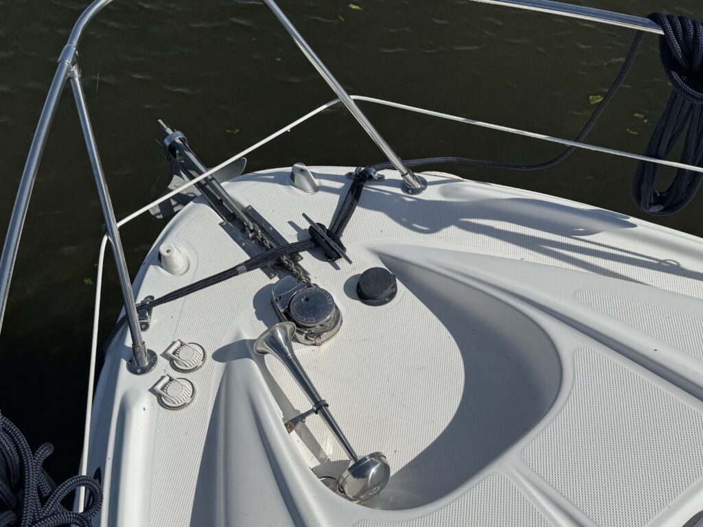 The bow of a white boat features a polished metal anchor winch with a rope guide. There are two metal cleats on either side and a circular hatch at the center, with an open area for the boat's deck on a body of water.