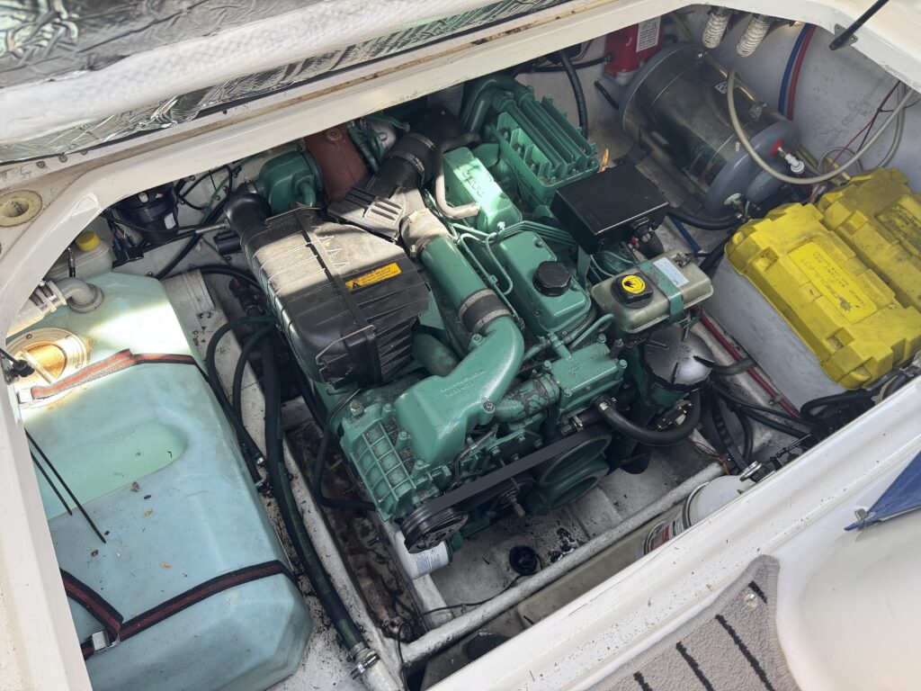 A green marine engine is visible in a compartment, surrounded by various components including hoses, wires, and a yellow battery. The compartment also contains a blue fuel tank and other equipment, all set within a white, enclosed space.