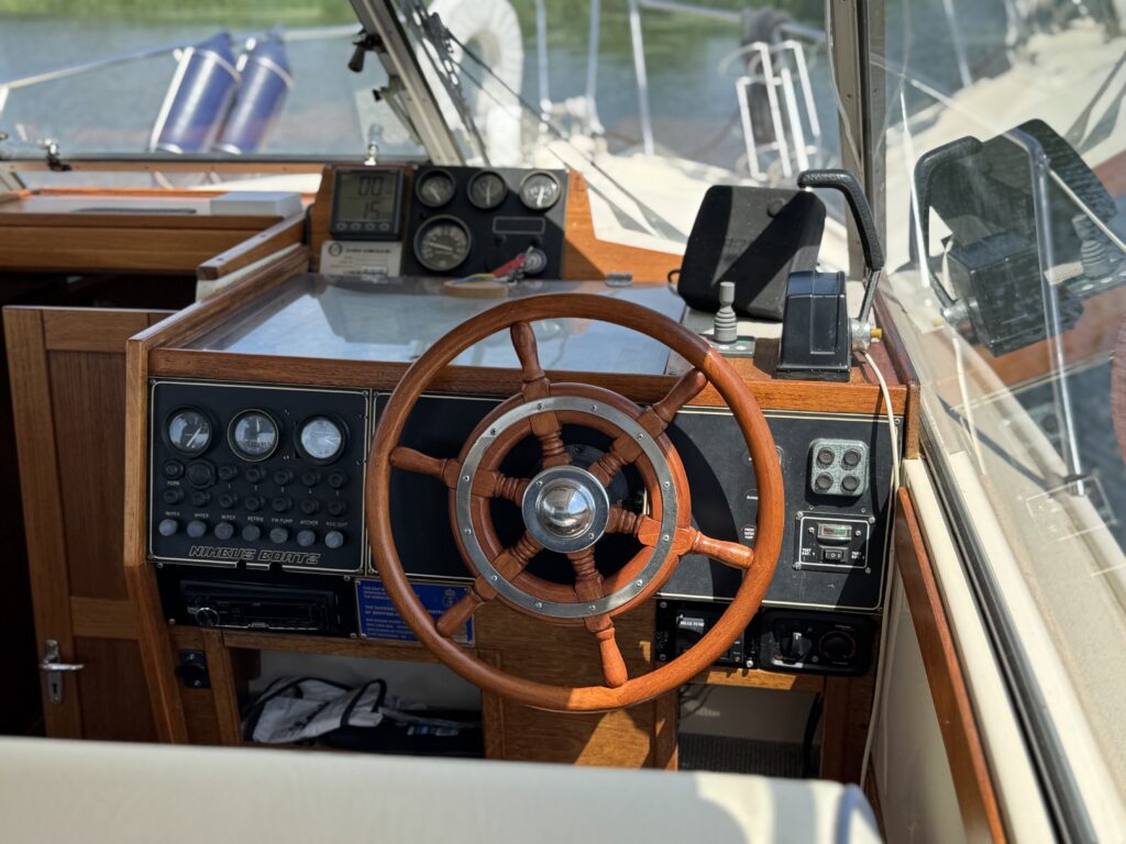 The interior of a boat's cockpit features a wooden steering wheel at the center with various control panels and instrumentation on either side. Natural light illuminates the space, highlighting a clean dashboard and sleek design.