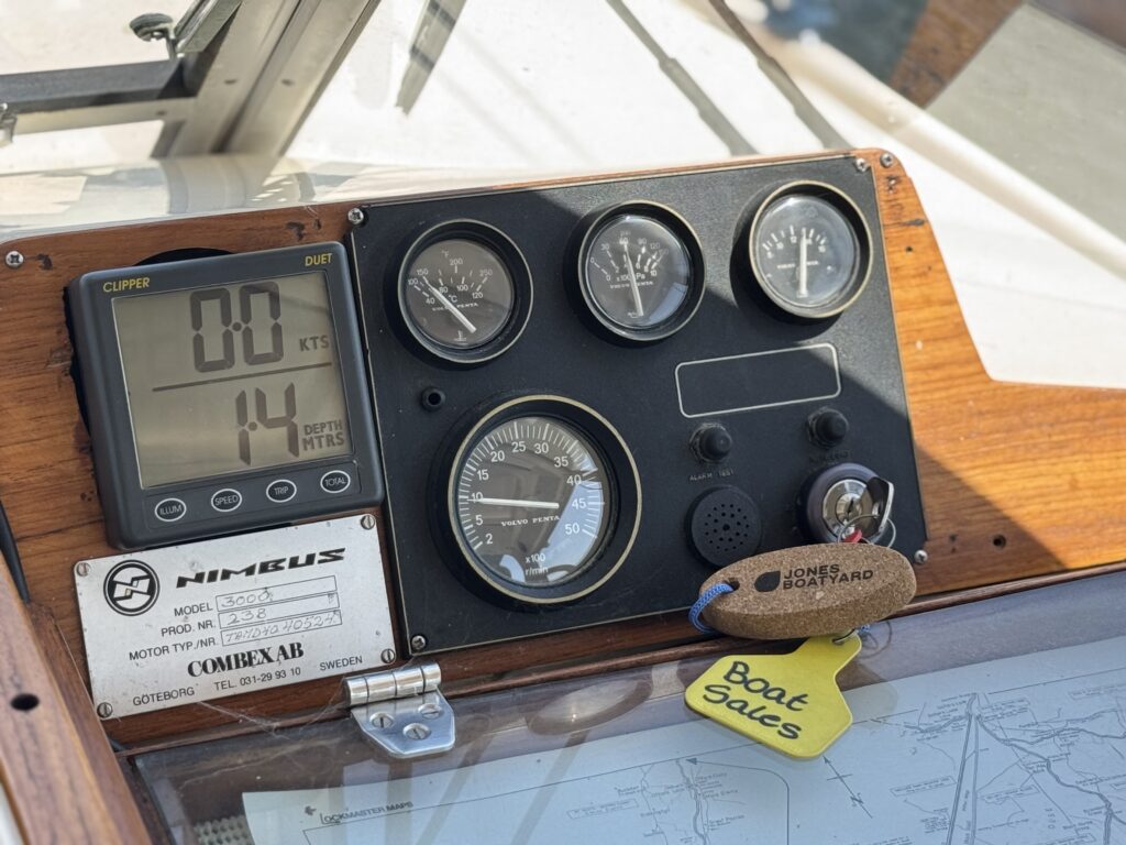A dashboard mounted on a boat displaying various instruments. The panel includes a digital speed display showing 14 knots, three analog gauges, and a clock, all set against a wooden background with a keychain labeled 'Boat Sales.'