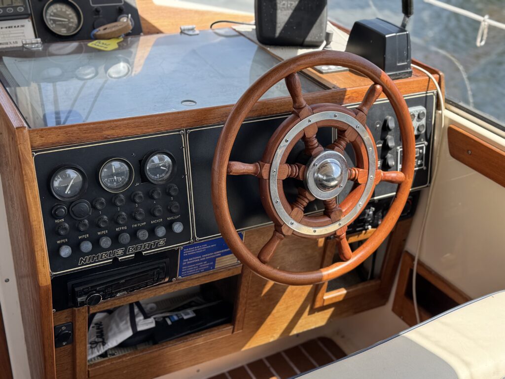 A wooden boat dashboard featuring a round steering wheel, various gauges, and control buttons. The panel is partially illuminated by natural light, and the console includes a radio and additional controls on the left side.