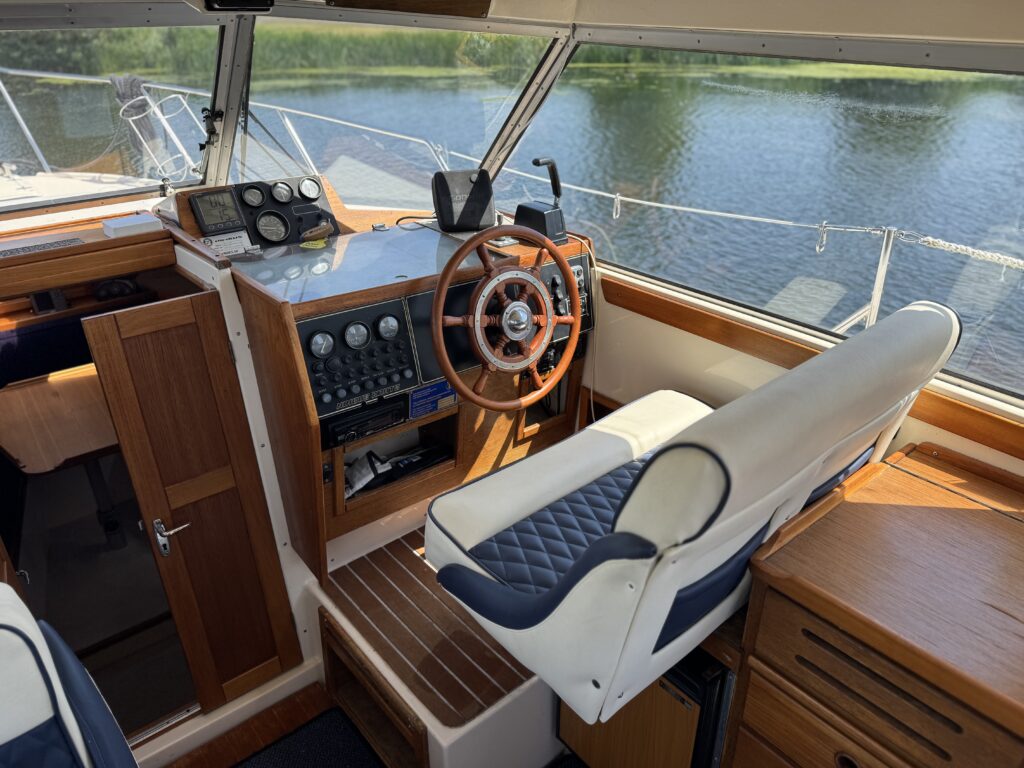 The interior of a boat's cockpit featuring a wooden steering wheel, control panels with various gauges, and a white leather captain's chair with blue accents. The background includes a view of water and greenery outside the windows.