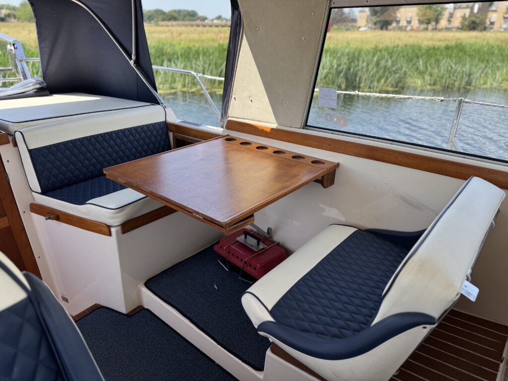 An interior seating area on a boat features two square upholstered benches with dark blue and cream-colored cushions. A wooden table sits between the benches, and a small red device is visible on the floor. Outside, green vegetation is reflected in the water.