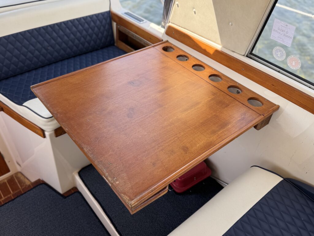 A wooden folding table mounted on a boat with a smooth surface and several cup holders along its edge. Dark blue, quilted cushions are visible on surrounding seats, and a window with natural light reflects off the table.