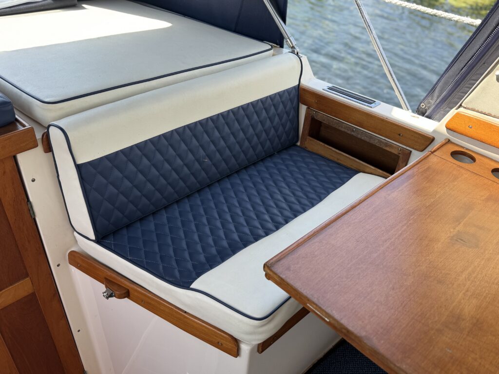 A covered boat seating area with a dark blue quilted cushion and light-colored edges. A wooden table is positioned nearby, with a smooth surface and cutouts for cup holders. The background shows water visible through a window.