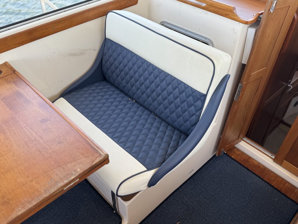 A cushioned bench seat with a navy blue and white upholstered design is positioned next to a wooden table in a boat interior. The surrounding area features wooden paneling and a textured blue carpet.