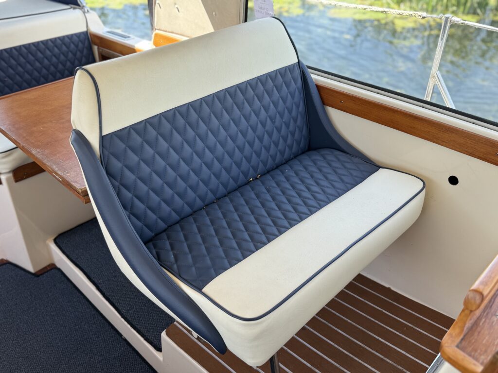 A boat interior features a blue and cream upholstered bench seat with diamond-pattern stitching. The seat is positioned near a wooden table and is surrounded by light streaming in from large windows, with water visible outside.
