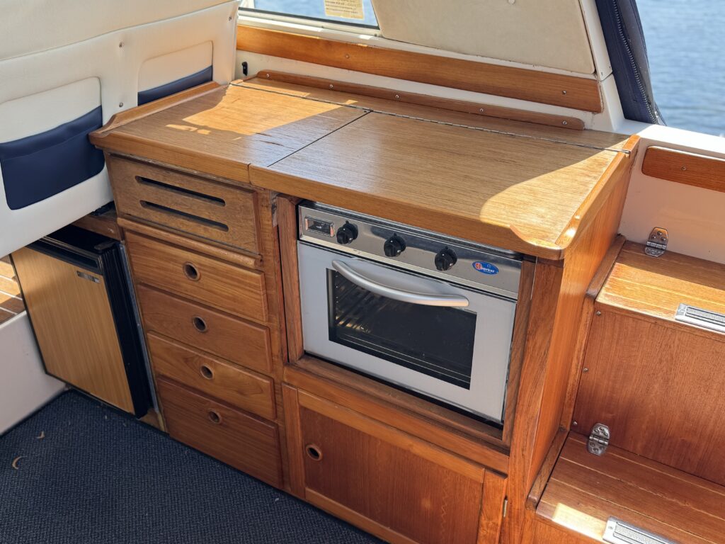 A wooden kitchen counter in a boat featuring a built-in oven with knobs, along with several drawers and cabinets for storage. The background shows a window with a view of the water and some natural light coming in.
