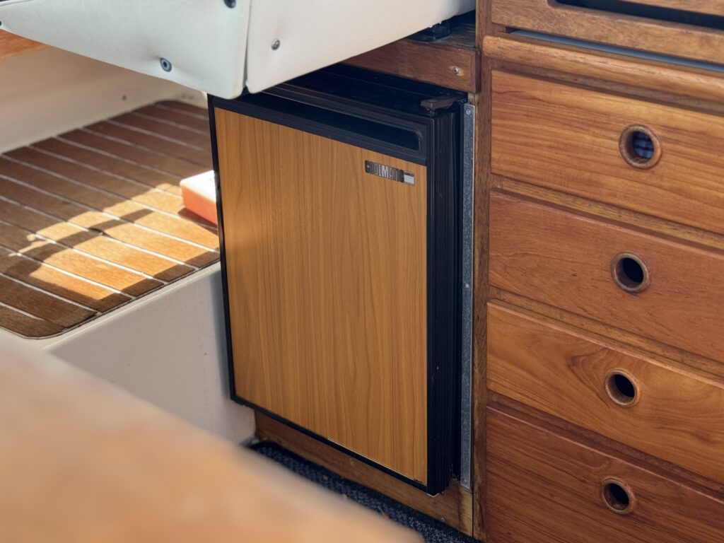 A compact refrigerator with a wooden panel front is installed in a boat's cabin. Nearby, there are wooden cabinets and a textured floor, creating a functional space for storage and appliances.