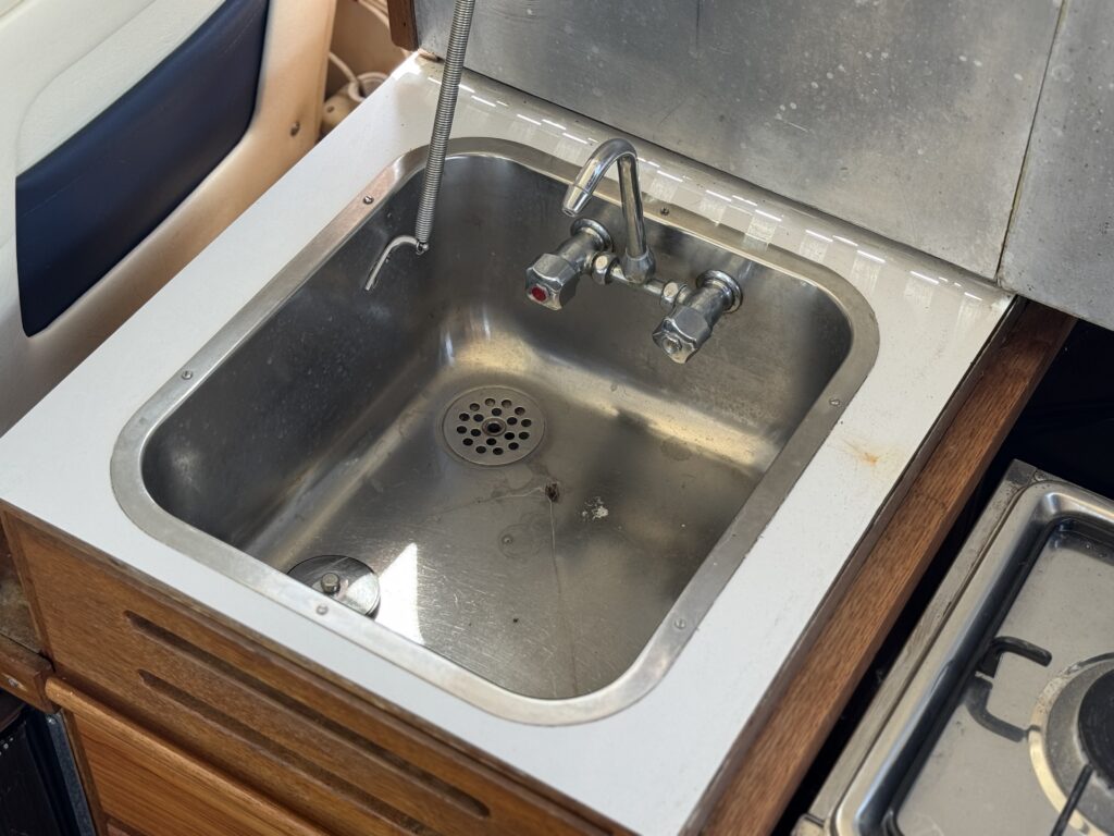 A stainless steel sink installed in a wooden countertop, featuring a single faucet with dual handles. The sink is empty and shows a drain at the bottom. The background includes part of a kitchen area with a stove and cabinetry.