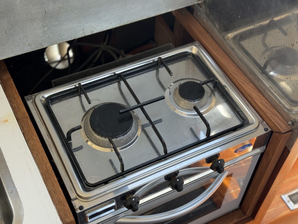 A stainless steel two-burner gas stove is installed in a wooden kitchen cabinet. The burners are round with a black surface, and the stove is equipped with control knobs at the front.