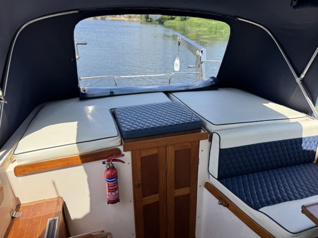 The interior of a boat featuring a cushioned seating area with white and blue upholstery. A wooden cabinet is situated beside a mounted fire extinguisher. In the background, natural light reflects off the water through an open window.