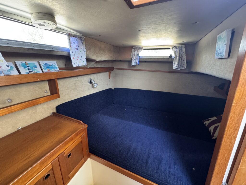 An interior cabin on a boat featuring a blue upholstered seating area that converts to a bed. The walls are adorned with light-colored panels and the space has wooden cabinetry and small windows with curtains, providing natural light.