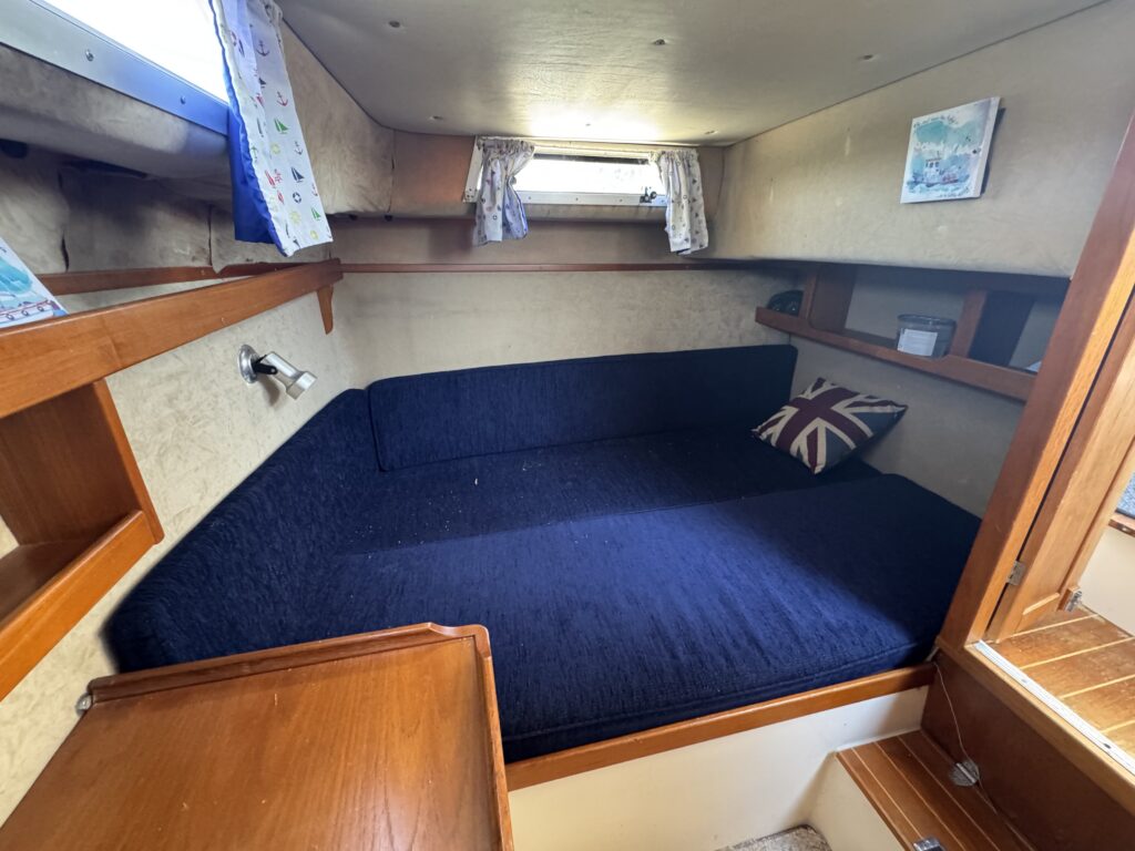 A cozy cabin interior features a dark blue cushioned seating area with a Union Jack pillow. Wooden shelves and a small table are visible, alongside windows with light curtains allowing natural light to enter.