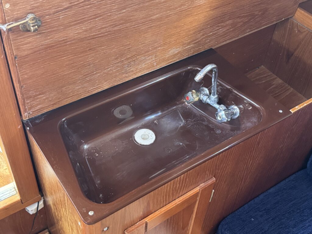 A brown sink made of plastic is installed in a wooden boat interior. The sink has a single faucet with a hot and cold water mixer, and a drain hole is visible at the bottom.