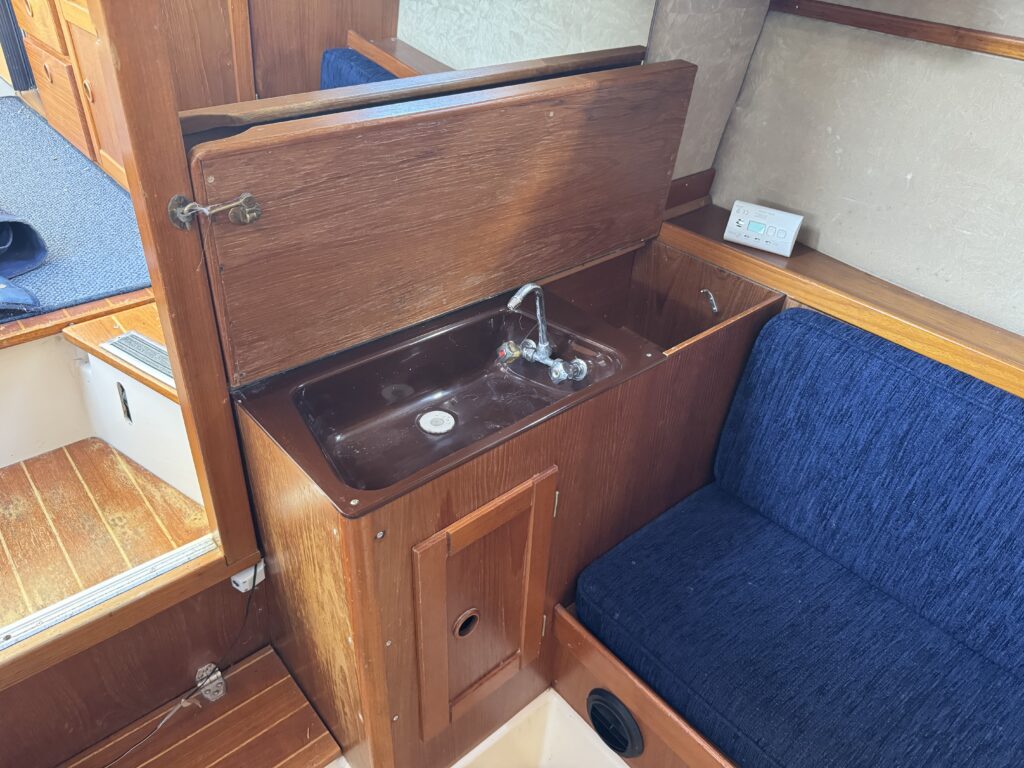 An interior view of a boat space featuring a brown wooden cabinet with a sink and faucet. Next to the sink is a small blue upholstered couch. The floor is made of wooden planks, and there are wooden cabinetry and a blue cloth visible in the background.