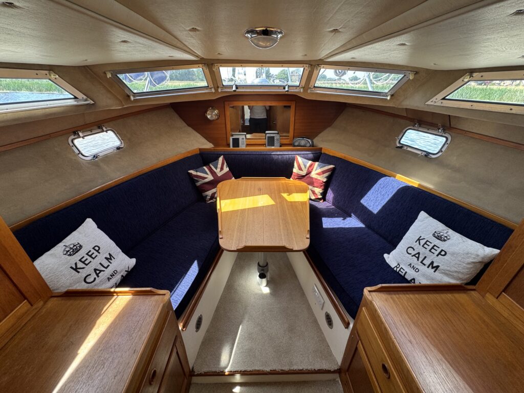The interior of a yacht featuring a seating area with a navy blue couch adorned with Union Jack cushions. A wooden table is centrally placed, and large windows allow natural light to fill the space.