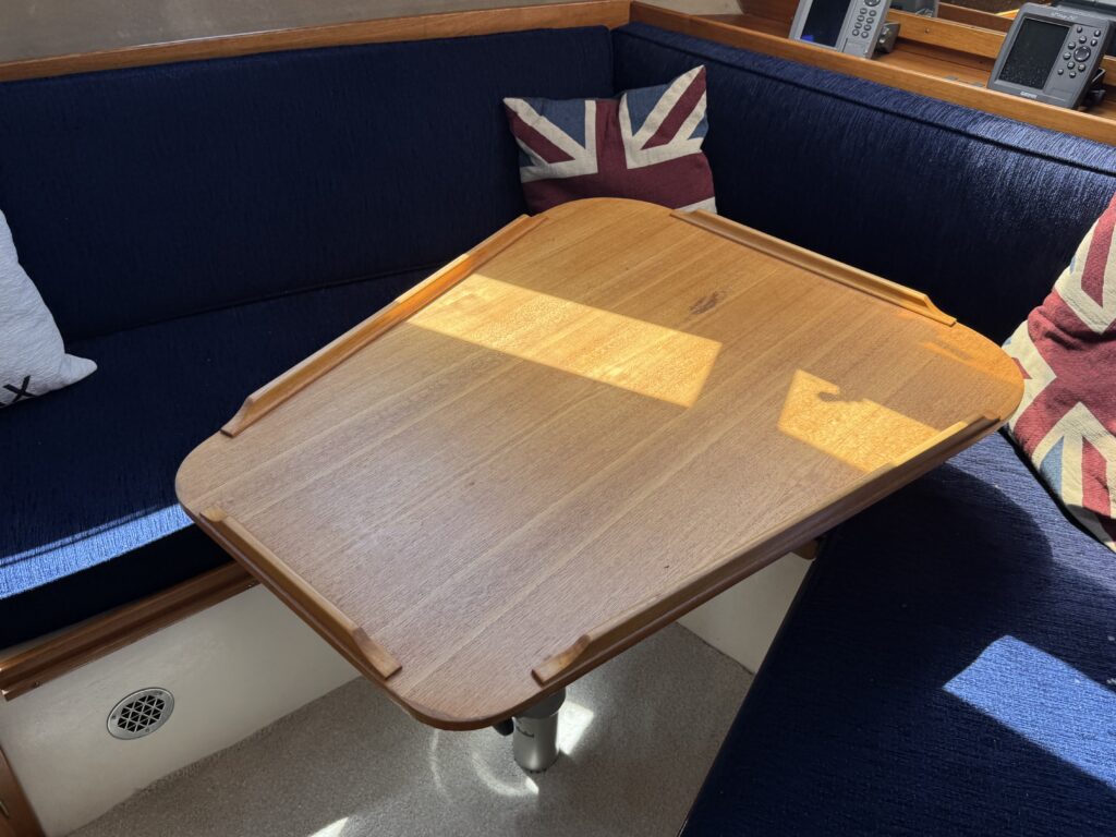 A rectangular wooden table positioned on a boat's interior. Surrounding the table are navy blue cushions, two of which have Union Jack designs. Sunlight is casting a warm glow on the table surface, highlighting its smooth finish.