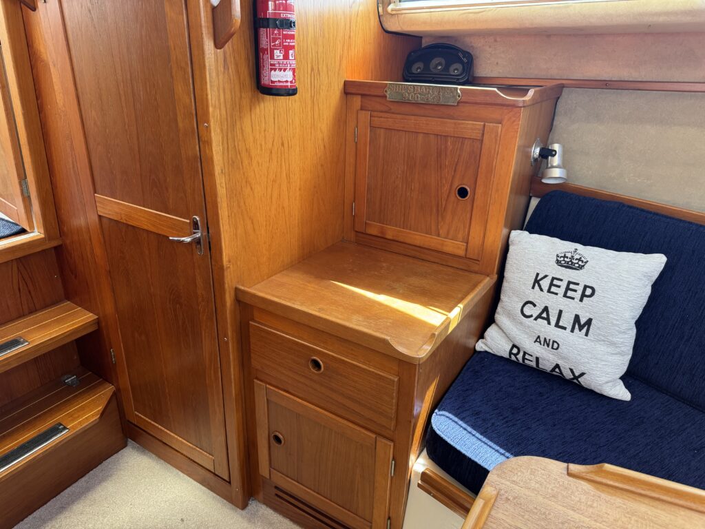 A cozy interior of a boat showing wooden cabinetry. A small wooden table sits next to a fitted couch with a cushion that reads "KEEP CALM AND RELAX." A closed cabinet is visible, along with a fire extinguisher mounted on the wall.