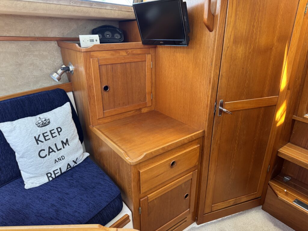 An interior view of a boat featuring a wooden cabinet and a bedside table. A blue cushioned seat with a decorative pillow that reads "KEEP CALM AND RELAX" is visible. A small television is mounted on the wall, and there are wooden paneling details throughout.
