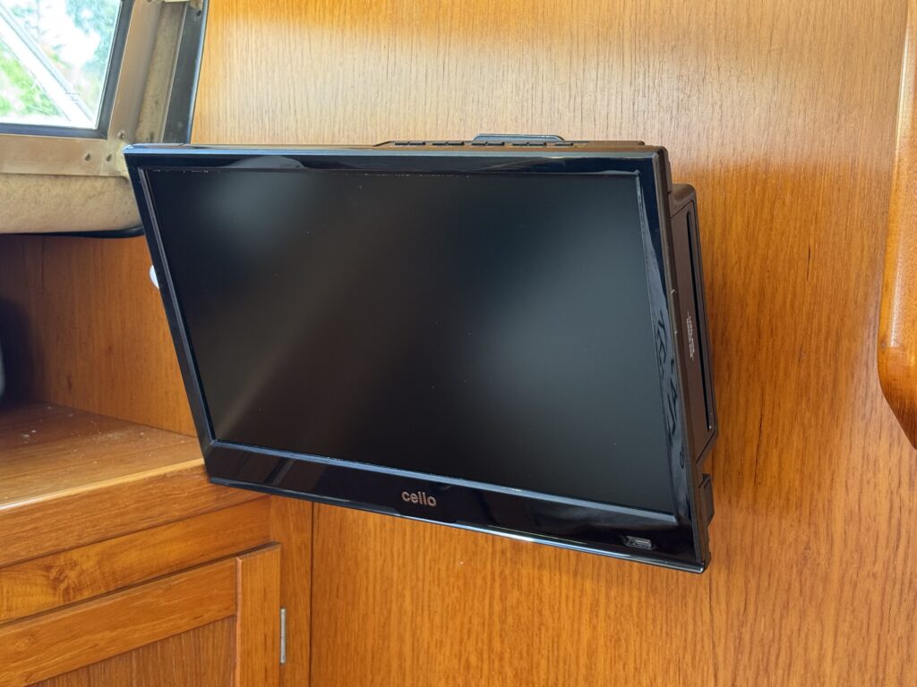 A flat-screen television mounted on a wooden wall in a boat interior. The TV is turned off, displaying a black screen, and nearby, a window allows natural light to illuminate the space.