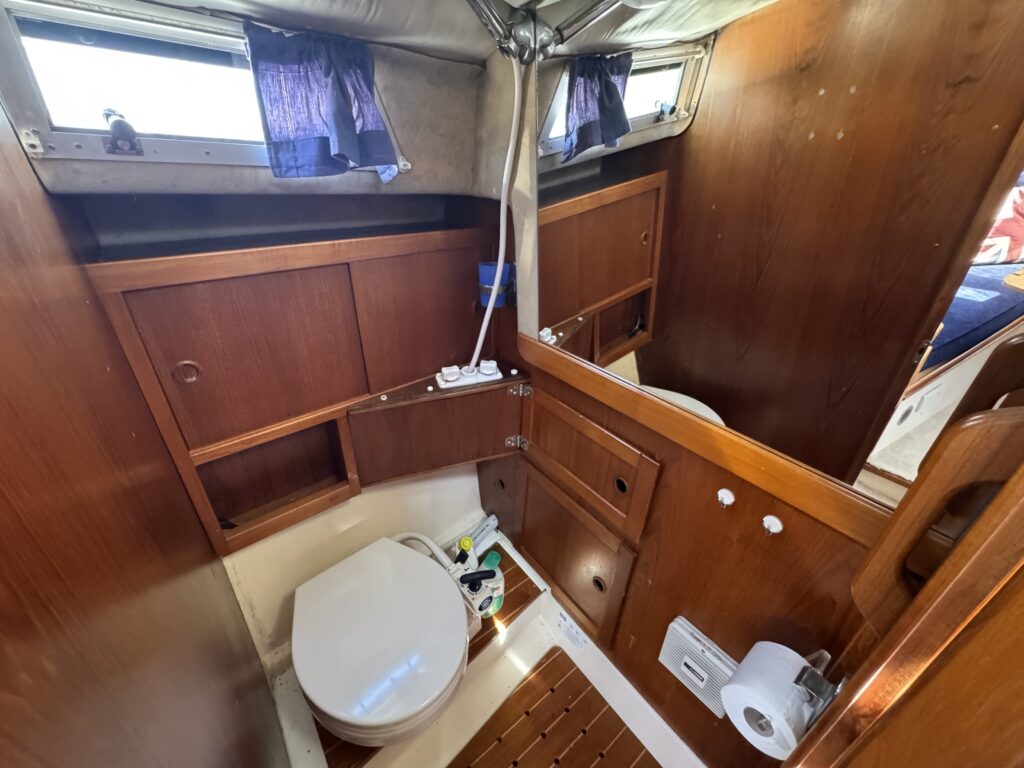 A compact bathroom on a boat featuring a white toilet, wooden cabinetry, and a small mirror. Natural light enters through windows covered with blue curtains. The floor has wooden slats, and there are toiletries placed near the toilet.