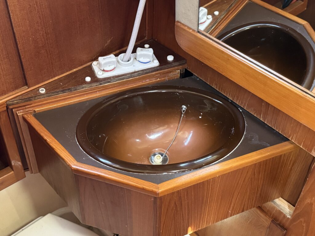 A brown oval sink with a chrome drain, set into a wooden vanity with an octagonal shape. Above, a mirrored panel reflects the sink, and electrical outlets are visible on the side, alongside two plumbing switches.
