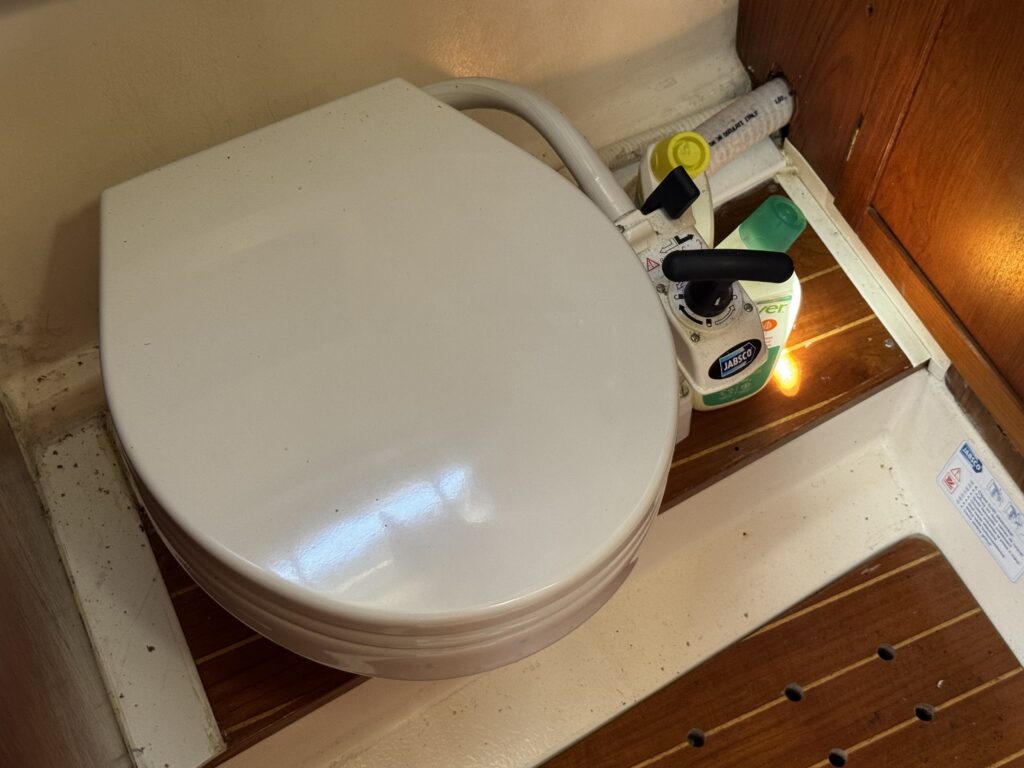 A white toilet with a closed lid is positioned next to a wooden step, with a small green cleaning bottle and a light source nearby. Visible plumbing components connect to the toilet, creating a functional installation in a compact space.