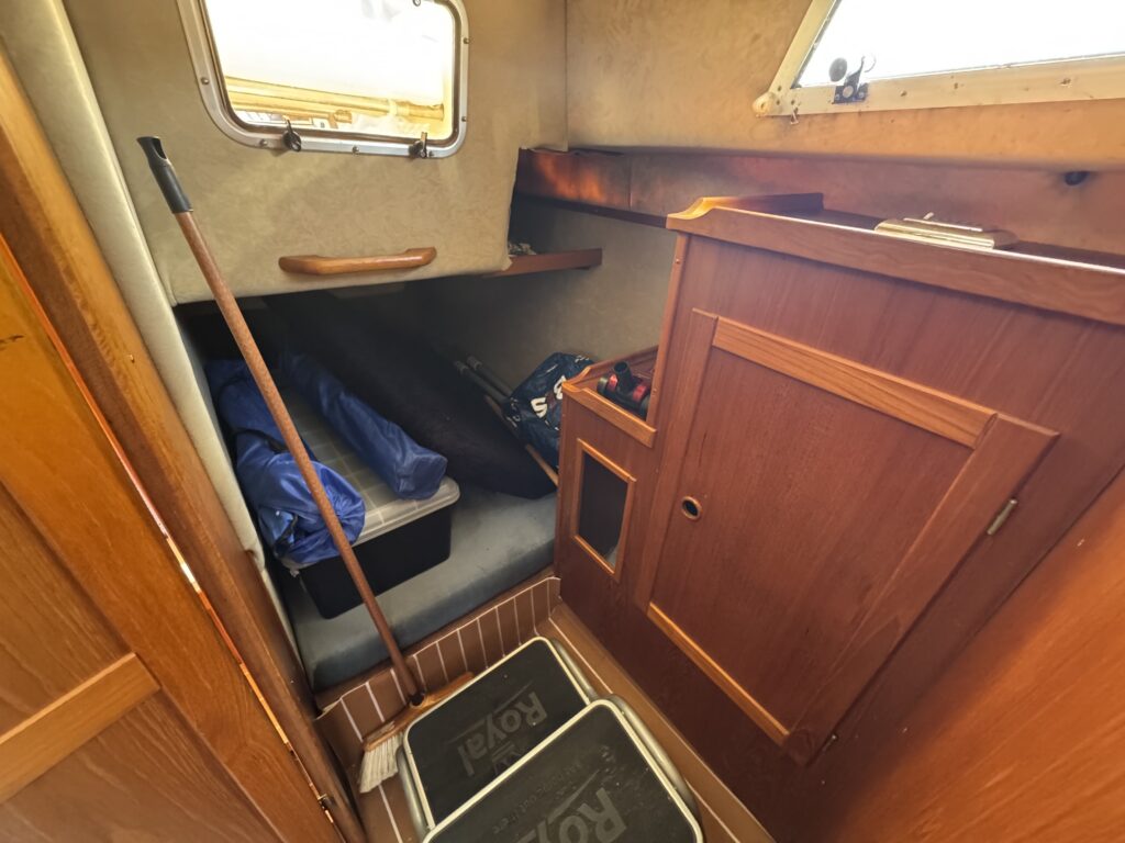 An interior boat space with wooden paneling, featuring a small bench and storage compartments. A blue bag and several items are placed on the bench. A wall-mounted window allows natural light to enter, and a broom is leaning against the wall.