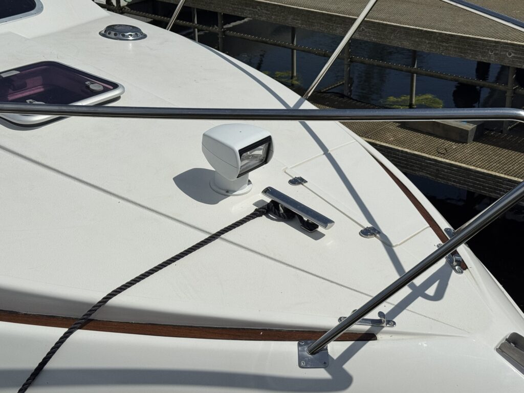 The deck of a boat featuring a white surface with a spotlight mounted, a stainless steel railing, and a cleat securing a black rope. In the background, a wooden dock is visible, reflecting in the water.
