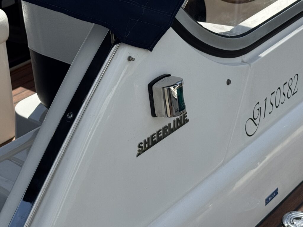 A close-up view of the side of a boat featuring the name "SHERLINE" in metallic lettering. A small metal detail is attached next to the name, and the background shows a portion of the boat's interior and seating area.